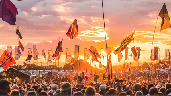 Glastonbury crowd at sunset, flags aloft including BBC 6 Music, Herne Bay, and Boyce banners.