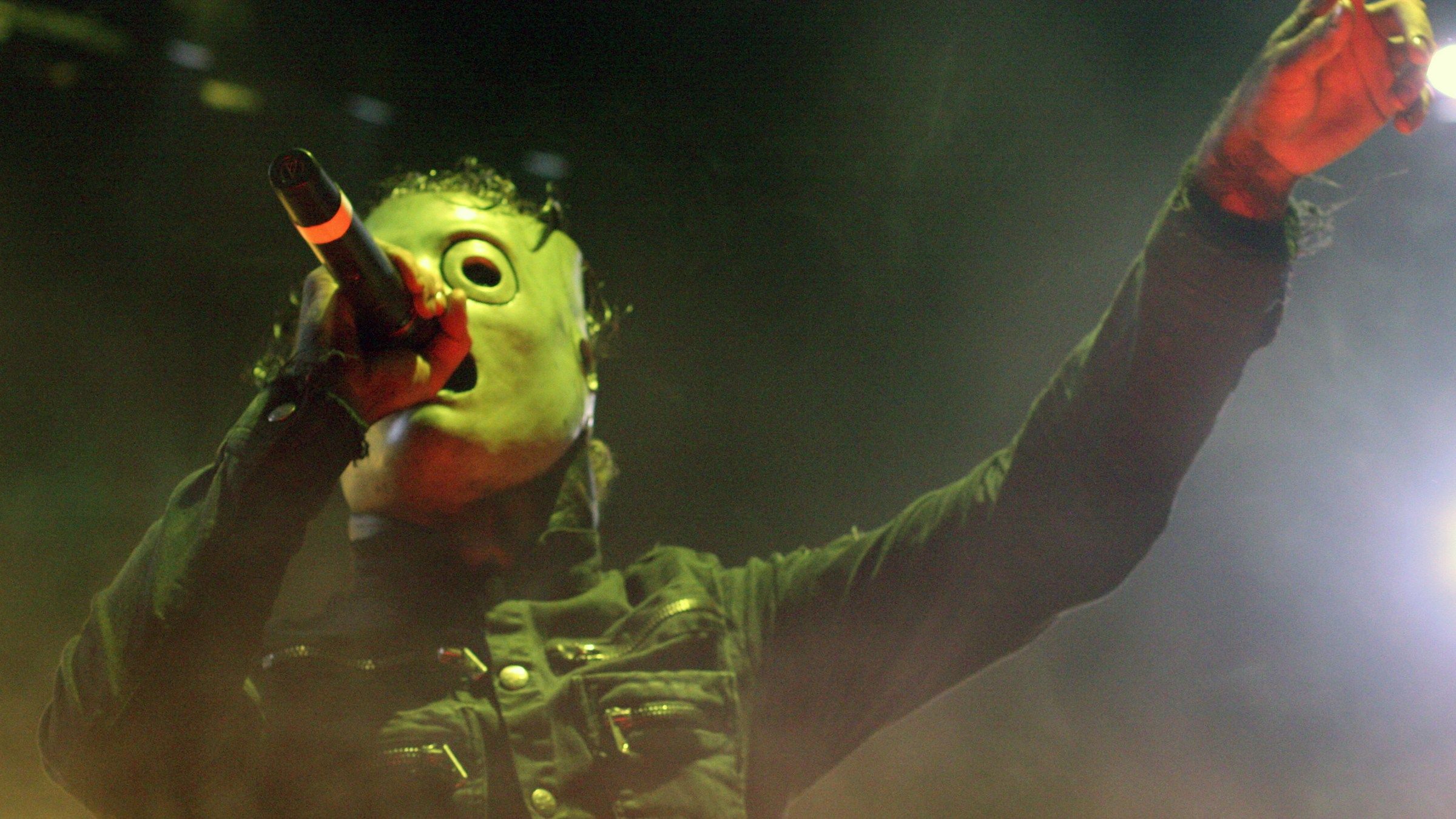 Corey Taylor of Slipknot singing in his mask with one arm stretched out, under green and gold stage light