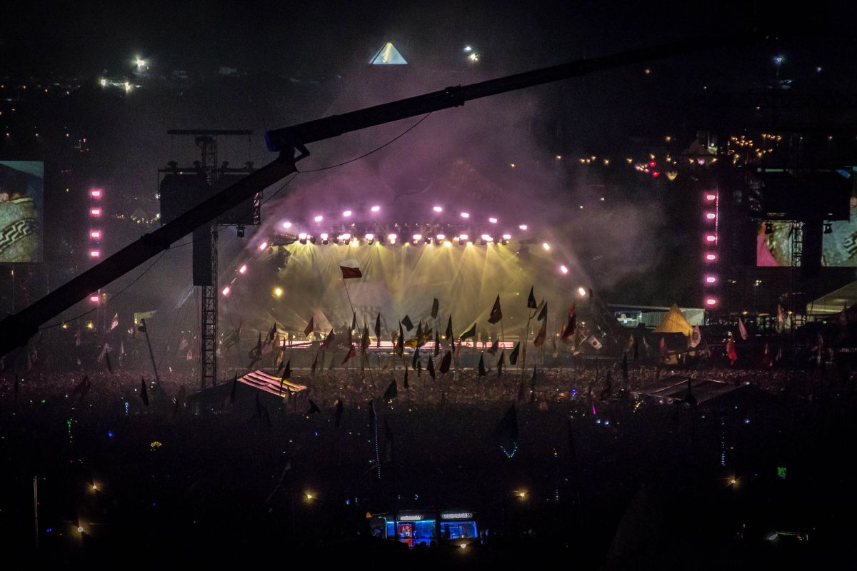 Night view of the lit Pyramid Stage with smoke and flags held up across a dark crowd