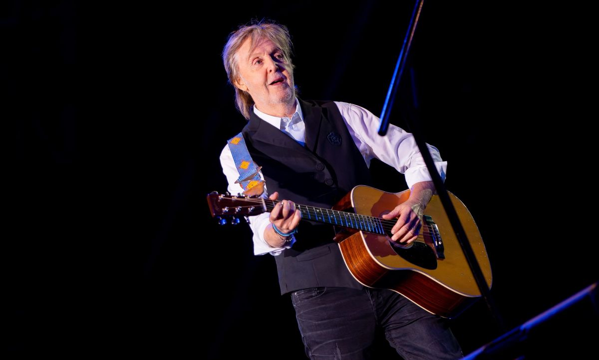 Paul McCartney in a white shirt and black waistcoat playing an acoustic guitar on a dark stage