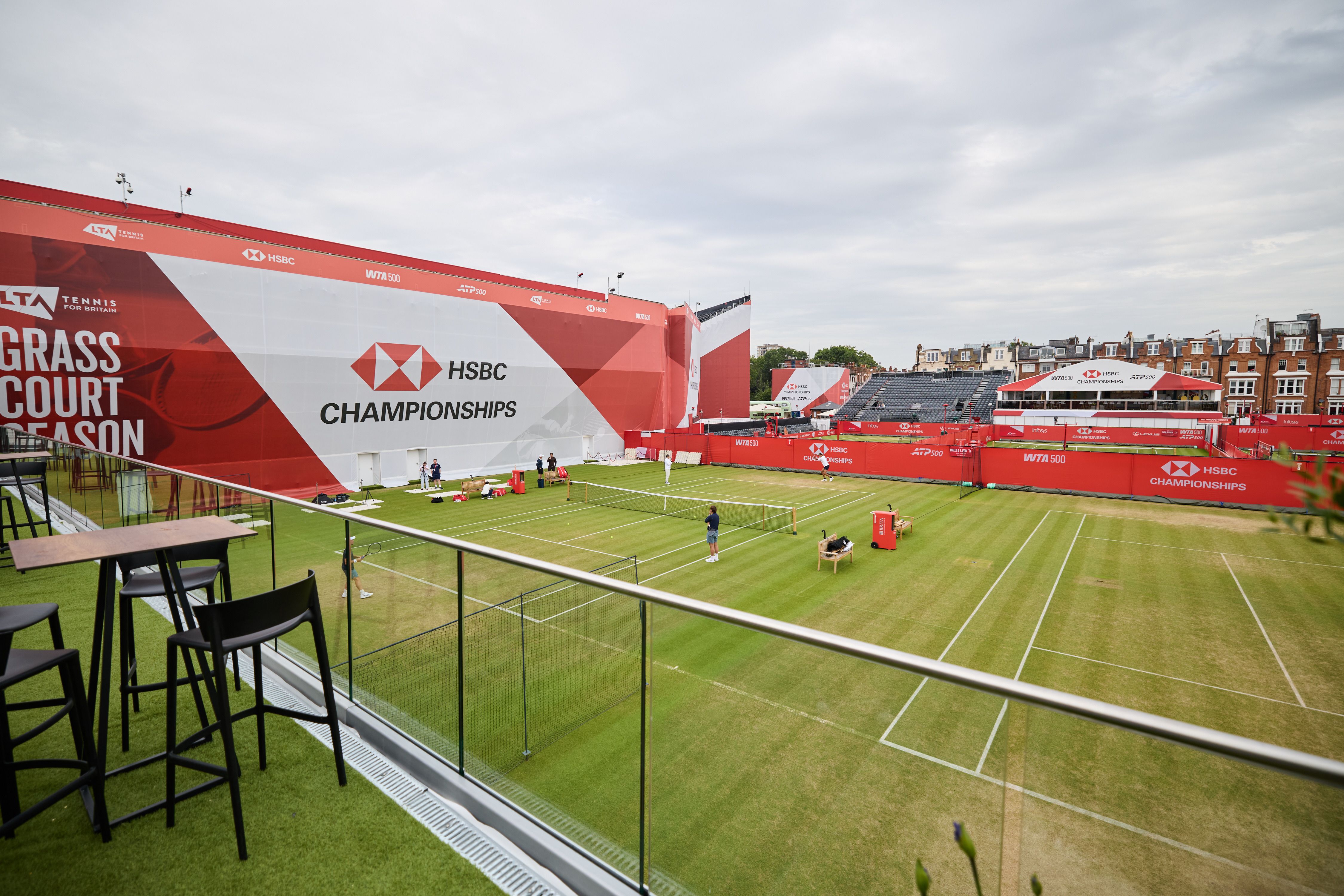 A hospitality terrace with high tables overlooking outer grass courts and HSBC branded boards