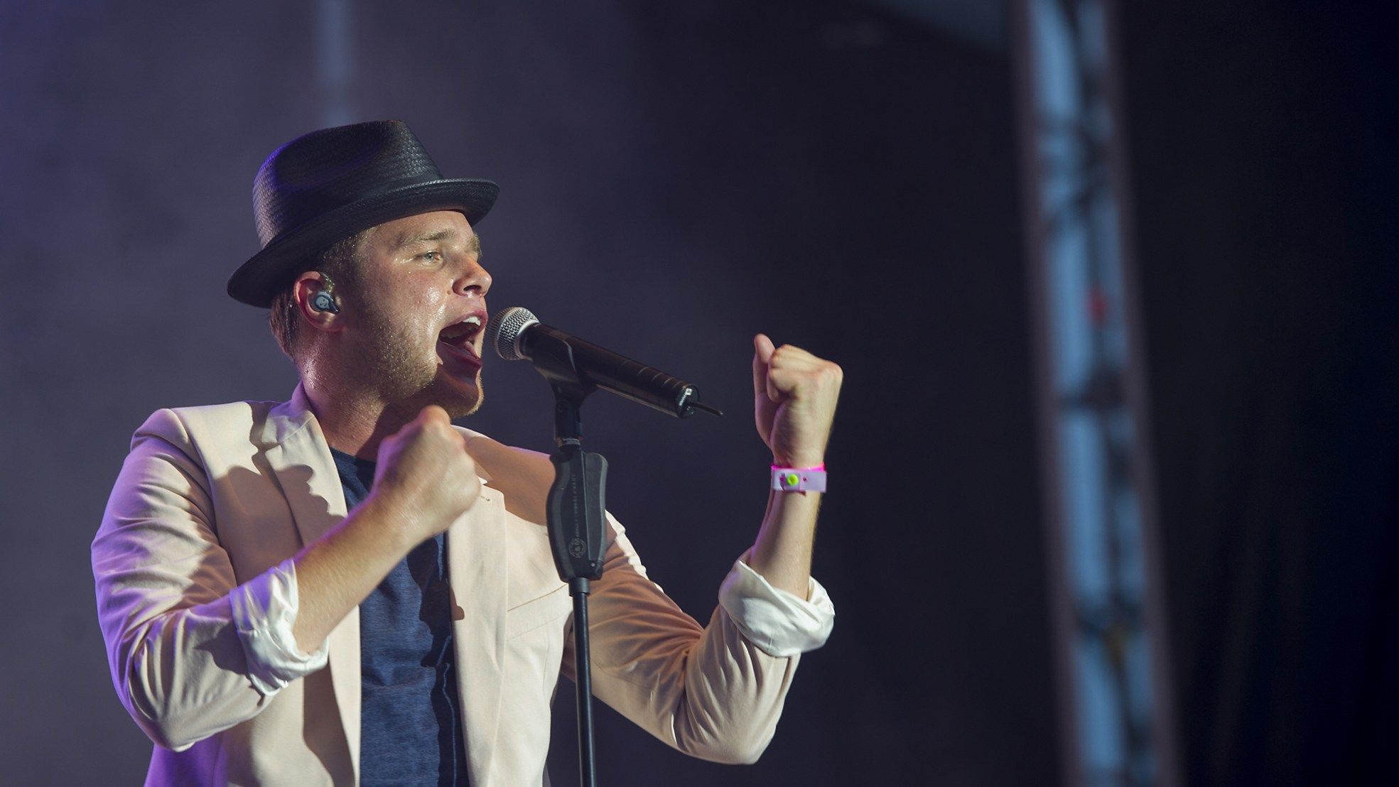 Olly Murs in a trilby and cream jacket singing into a microphone stand with his fists raised