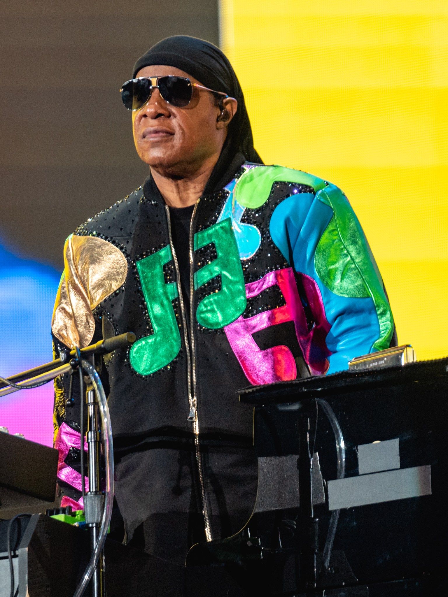 Stevie Wonder standing at his keyboards on stage in a black jacket covered in coloured music notes
