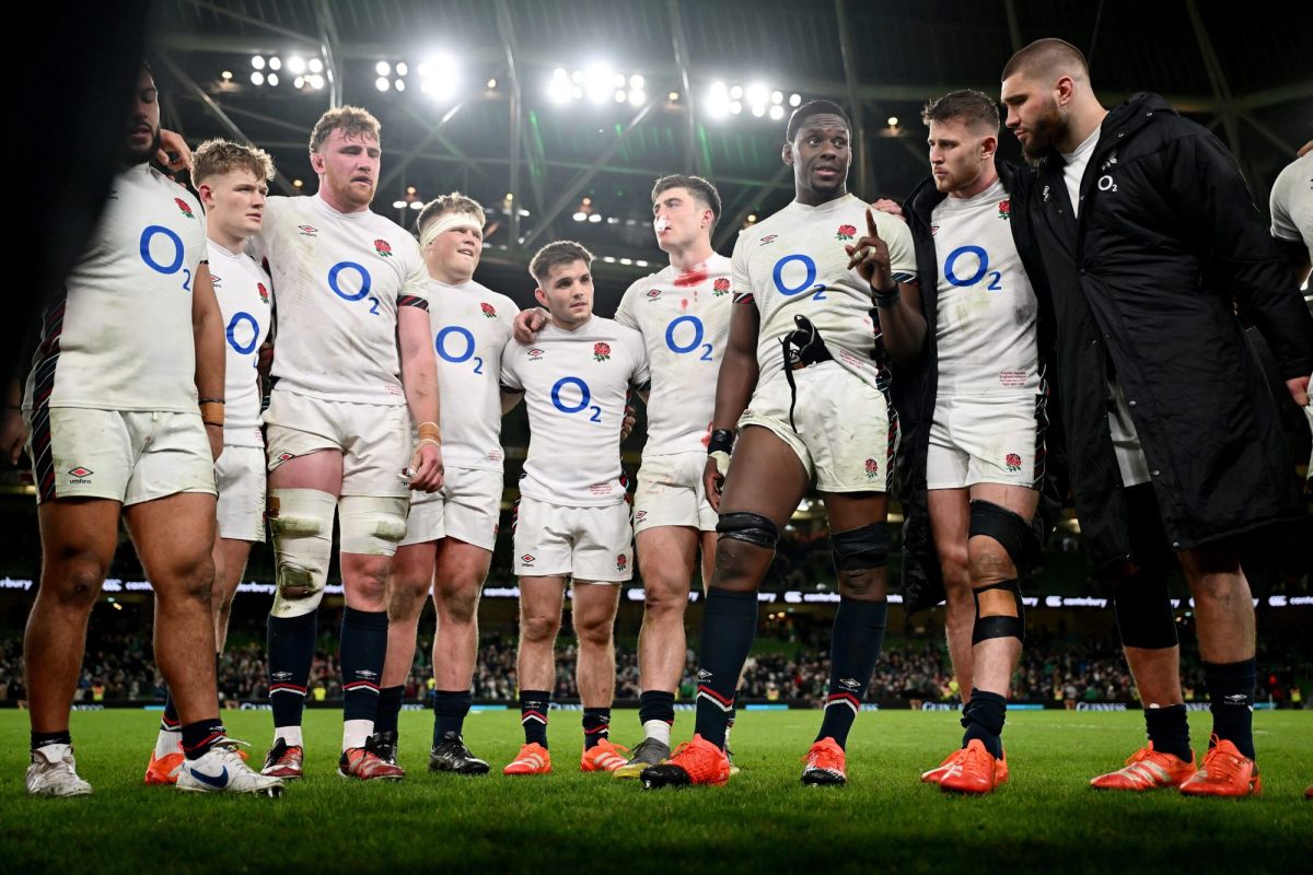 England rugby players in their white O2 sponsored kit huddle on the pitch during a Six Nations match, with stadium light