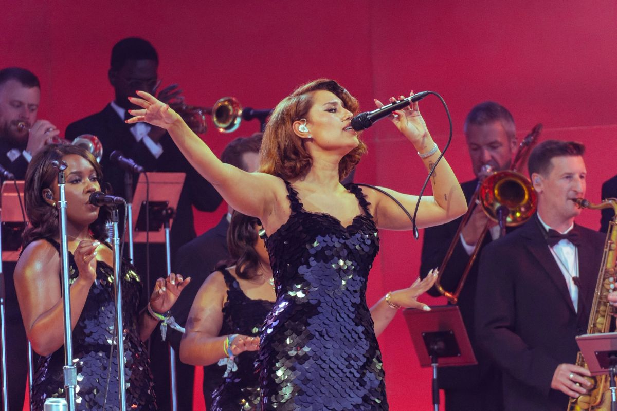 RAYE singing into a handheld microphone in a sequinned dress, arm outstretched, with a brass section behind her