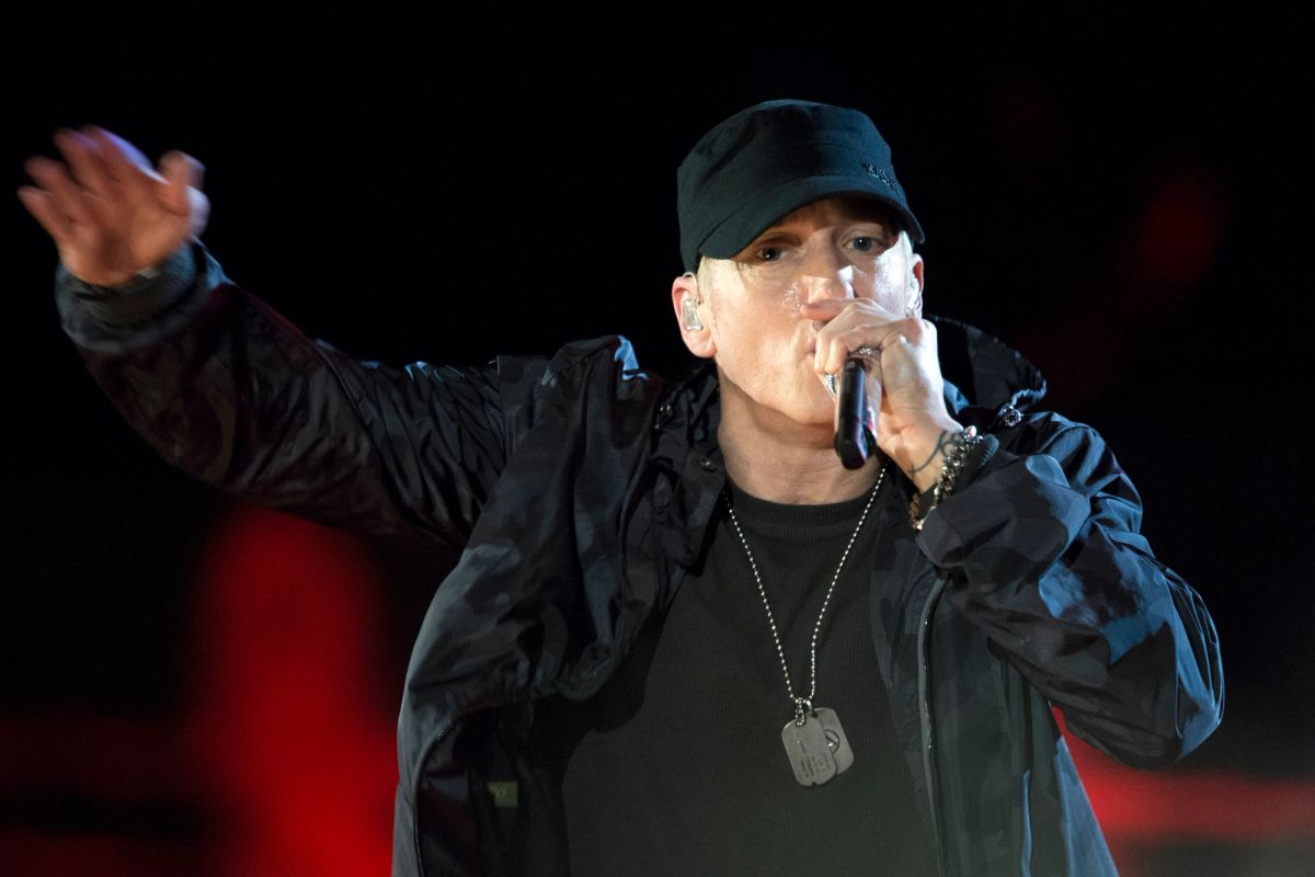 Eminem in a black cap and jacket rapping into a microphone with one arm stretched out