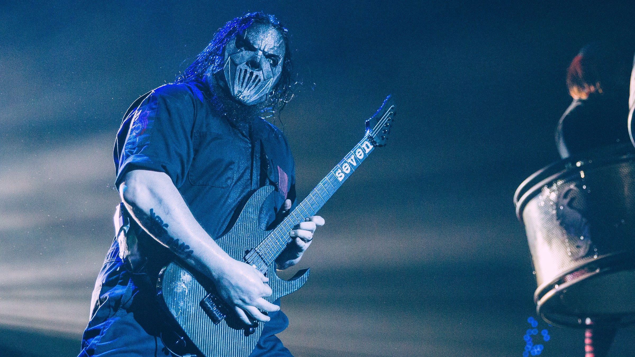 Slipknot guitarist Mick Thomson in his mask playing a seven-string guitar on stage under blue light