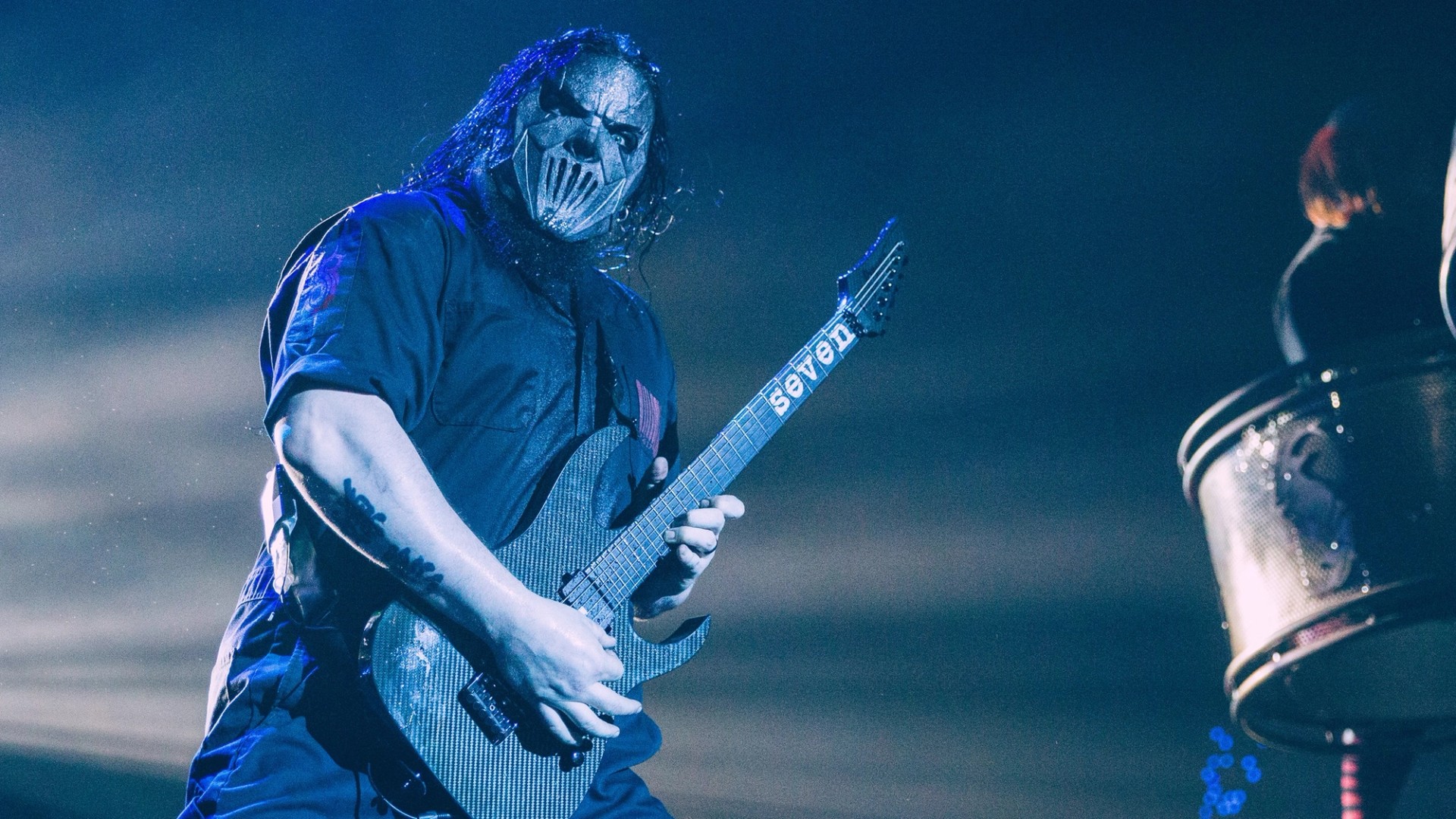 Slipknot guitarist Mick Thomson in his mask playing a seven-string guitar on stage under blue light