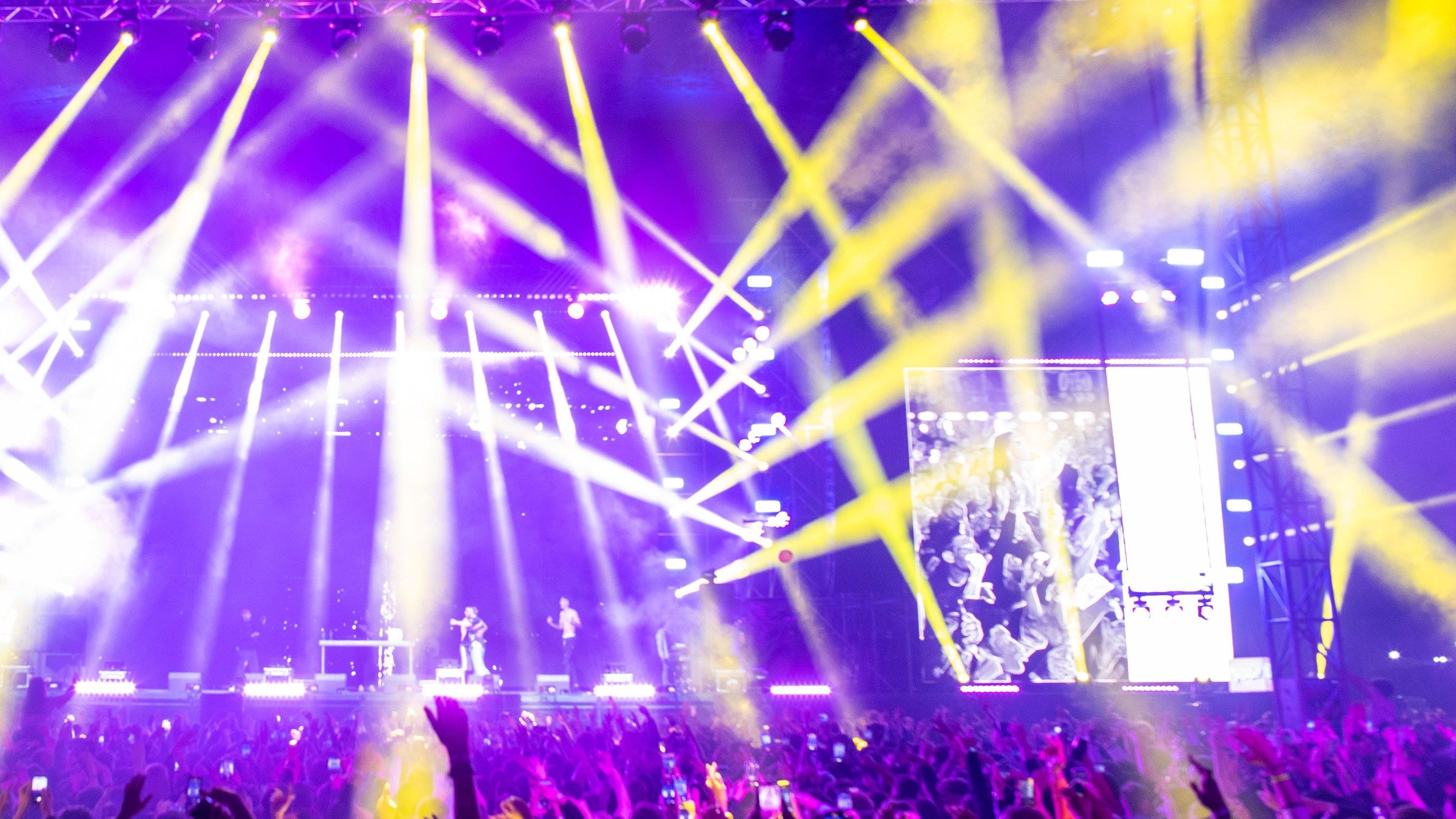 A crowd with raised hands and phones under sweeping purple and yellow stage light beams at a night concert