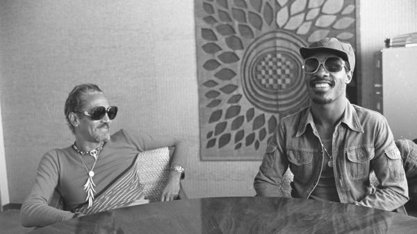 Stevie Wonder, smiling in large sunglasses and a cap, sitting at a table with music executive Ewart Abner in Los Angeles in 1975
