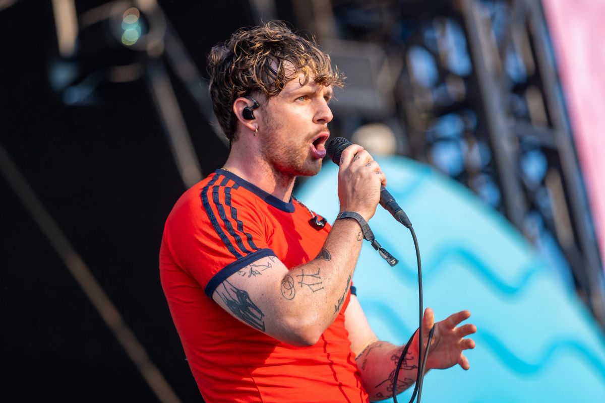Tom Grennan singing into a microphone on stage, wearing a red football-style shirt
