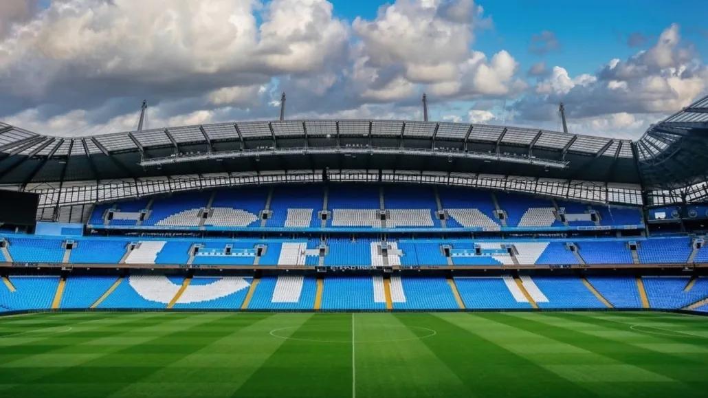 Wide-angle view of Etihad Stadium pitch with bold CITY lettering in sky-blue seats under dramatic clouds