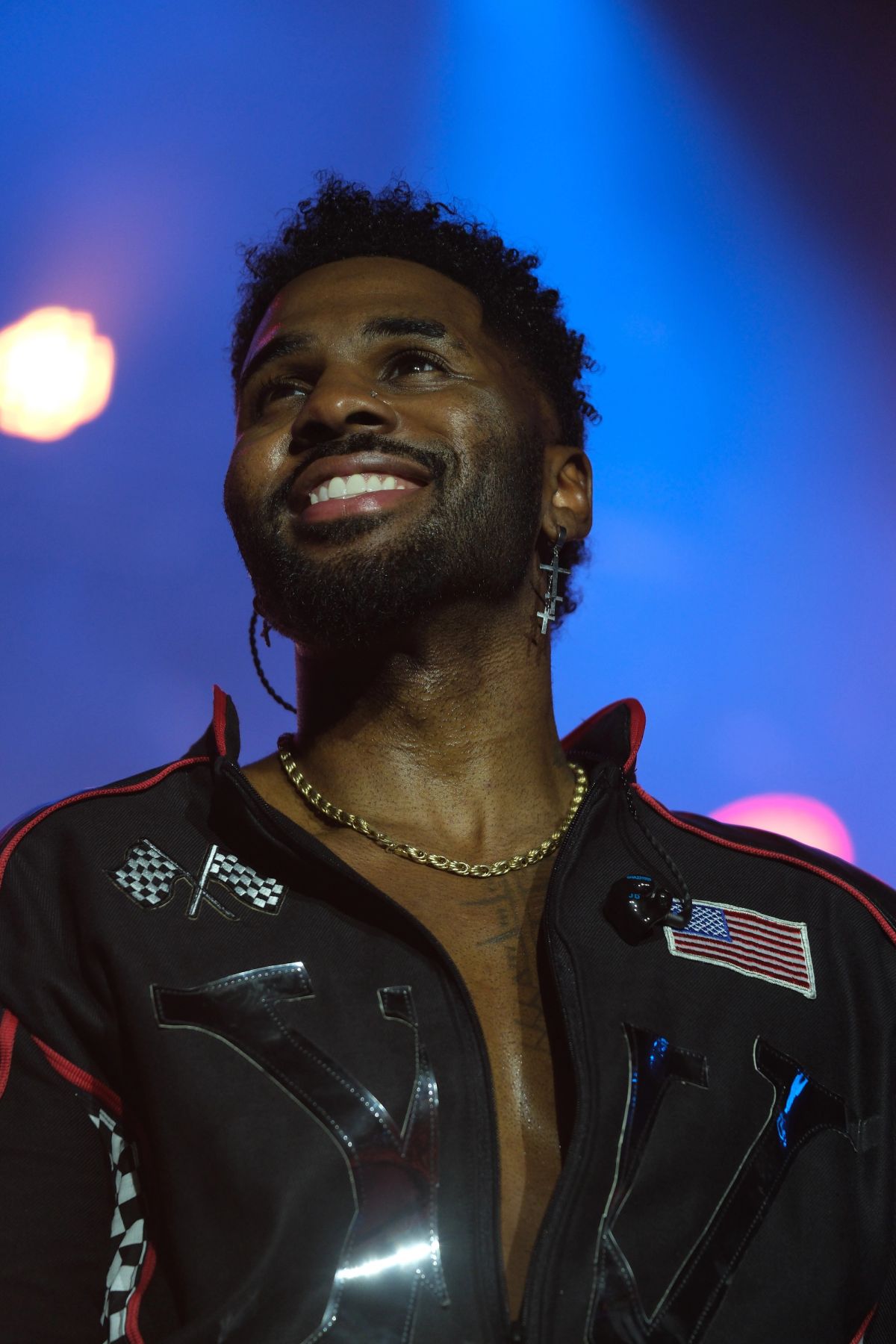 Jason Derulo smiling on stage in a black racing-style jacket under blue lights