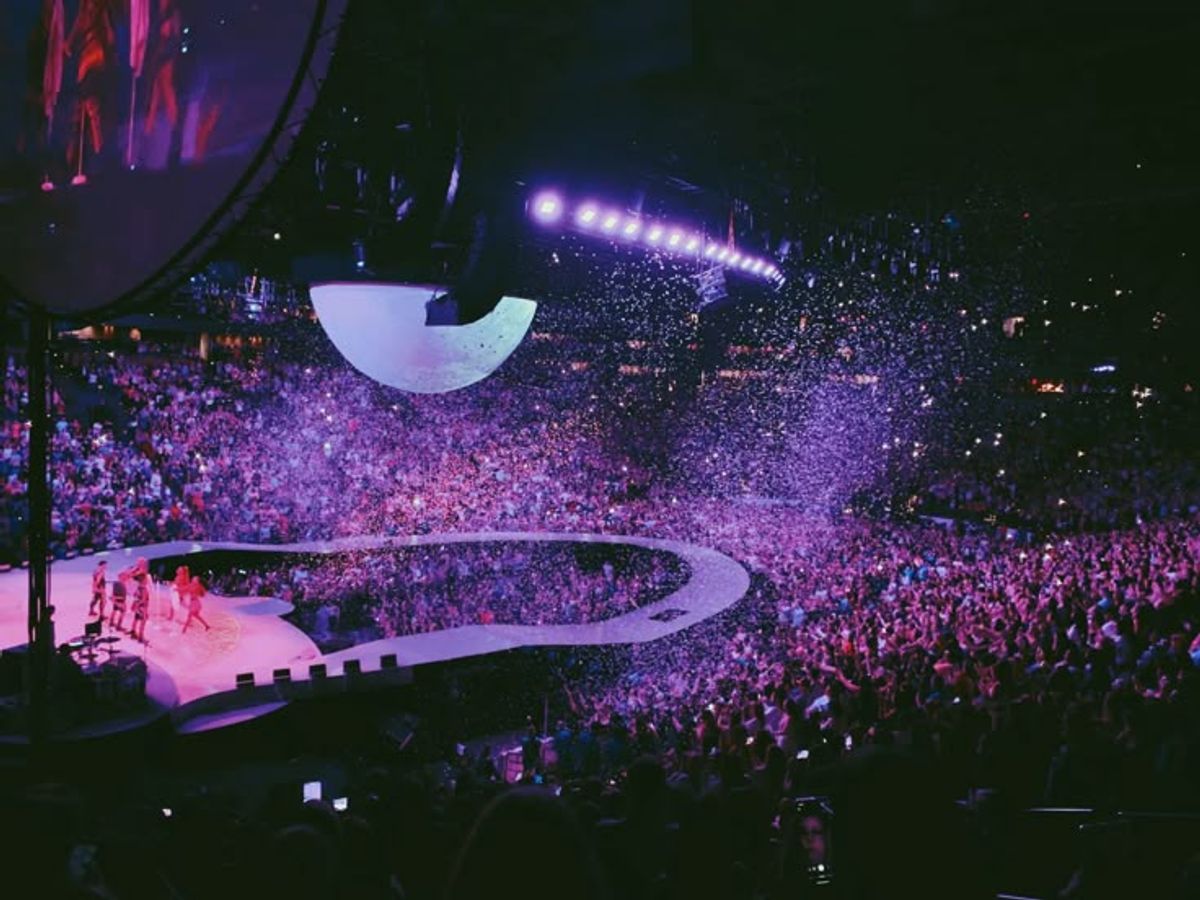 Dynamic arena concert with performers on a curvy stage under a crescent moon, as purple light bathes cheering crowd...