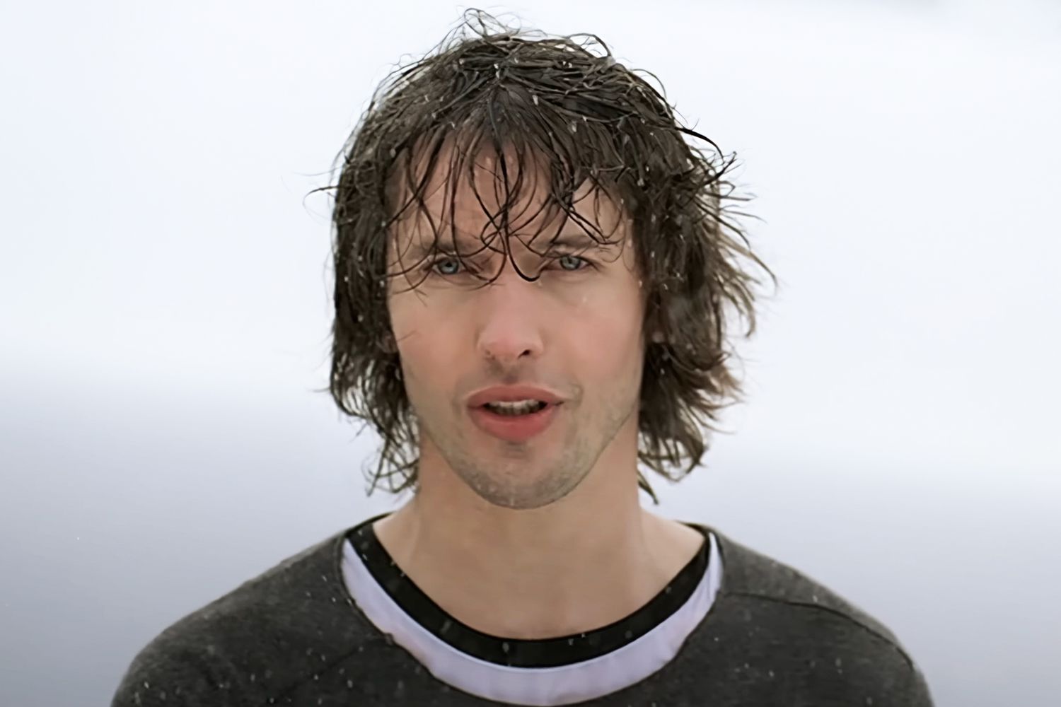 James Blunt with long wet hair singing in the rain against a pale grey sky, wearing a dark top