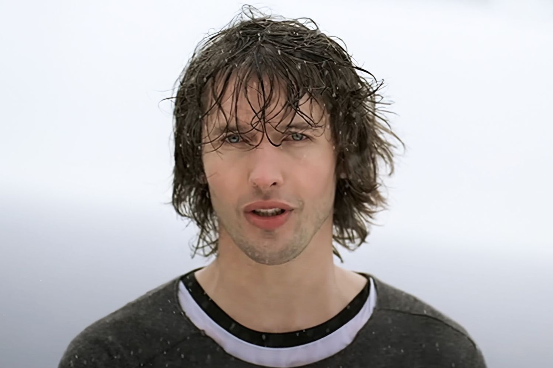 James Blunt with long wet hair singing in the rain against a pale grey sky, wearing a dark top