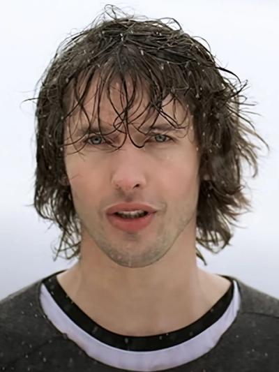 James Blunt with long wet hair singing in the rain against a pale grey sky, wearing a dark top