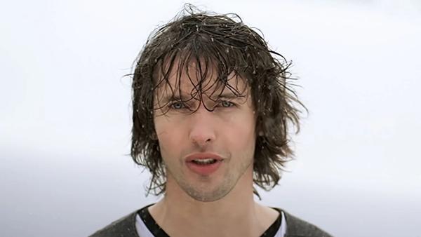 James Blunt with long wet hair singing in the rain against a pale grey sky, wearing a dark top