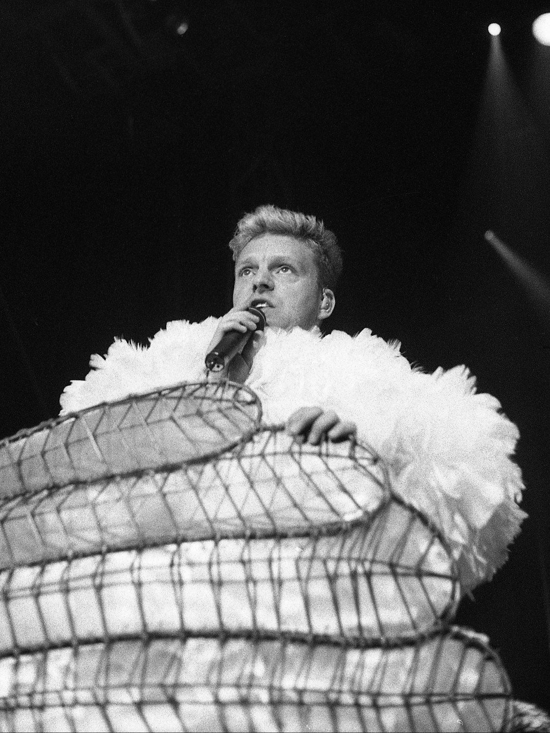 Black and white photo of Andy Bell of Erasure singing on stage, surrounded by white feathers