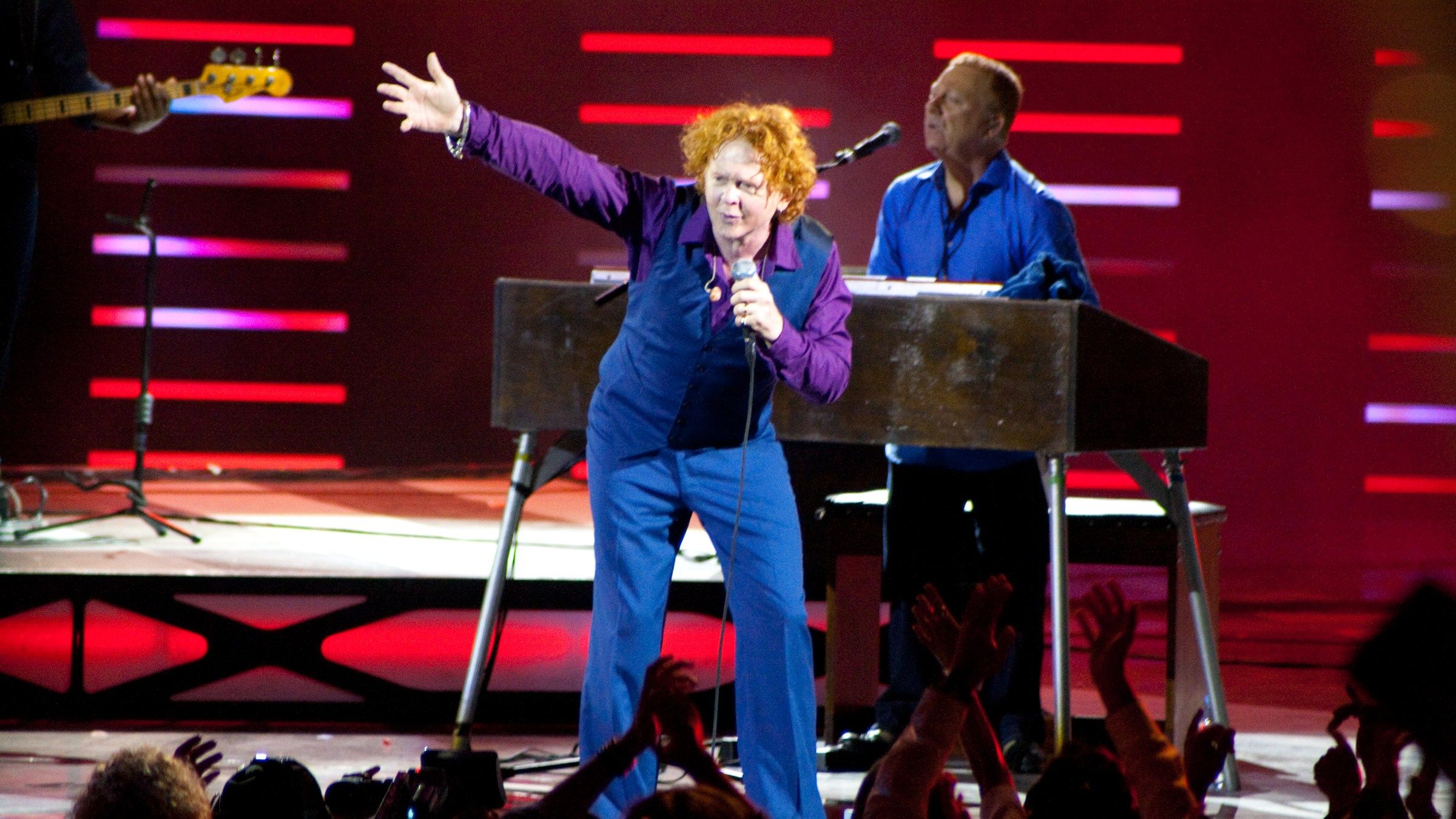 Mick Hucknall of Simply Red, in a blue suit and purple shirt, reaching out to the crowd on stage with the keyboard player behind him