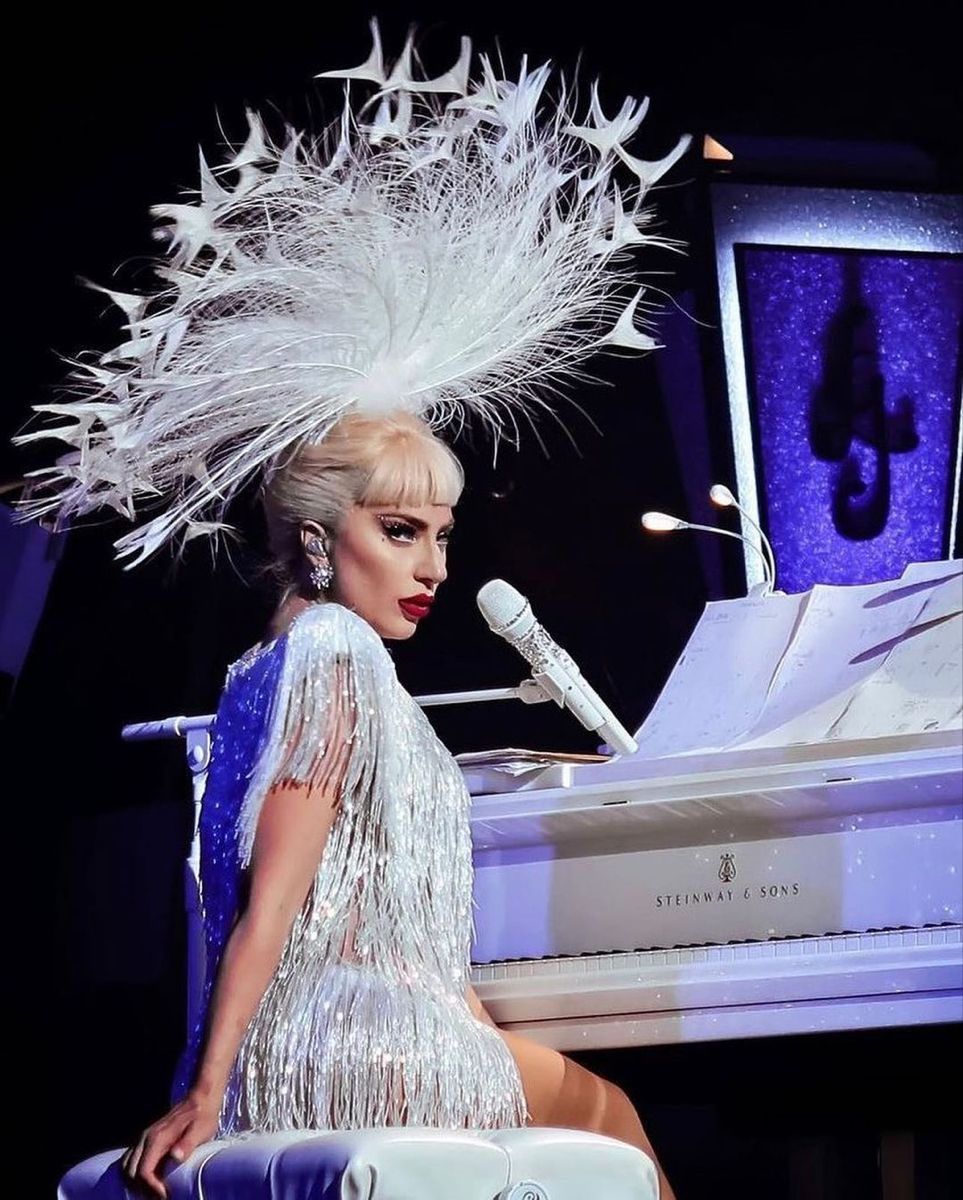 Lady Gaga, in silver fringe and feather headdress, sings at a white Steinway grand piano.