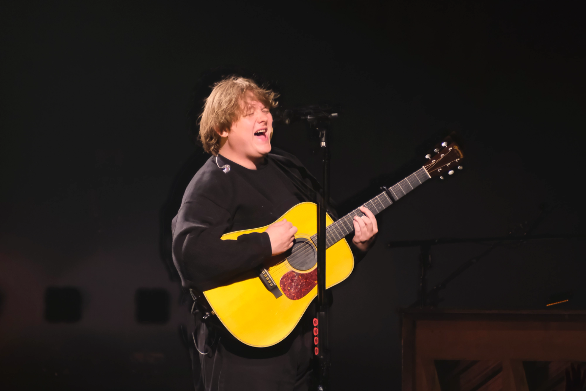 Lewis Capaldi singing and playing a yellow acoustic guitar on a dark stage