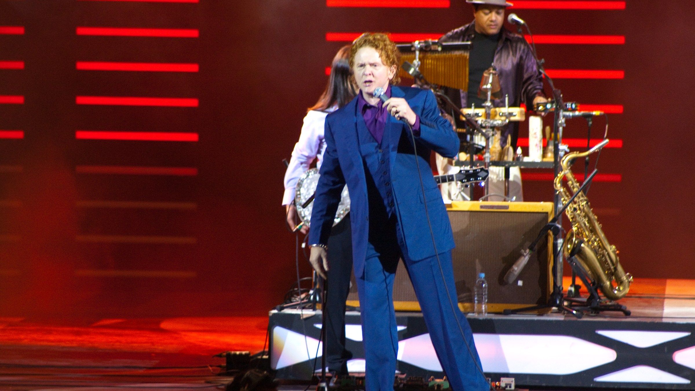 Mick Hucknall in a blue suit singing on a red-lit stage, with a percussionist and a saxophone behind him