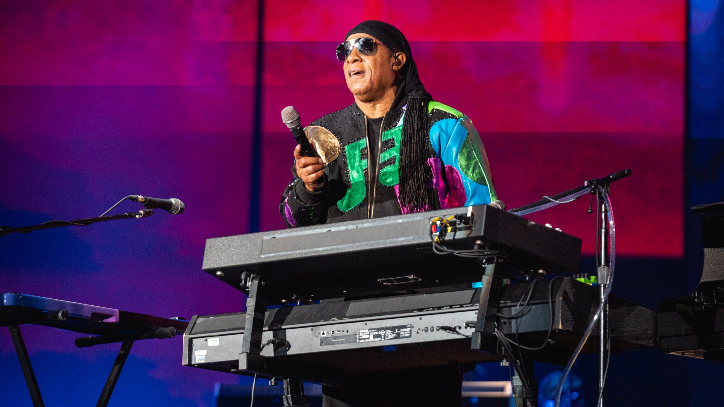 Stevie Wonder singing into a microphone behind his keyboard, with pink and purple stage lighting behind him