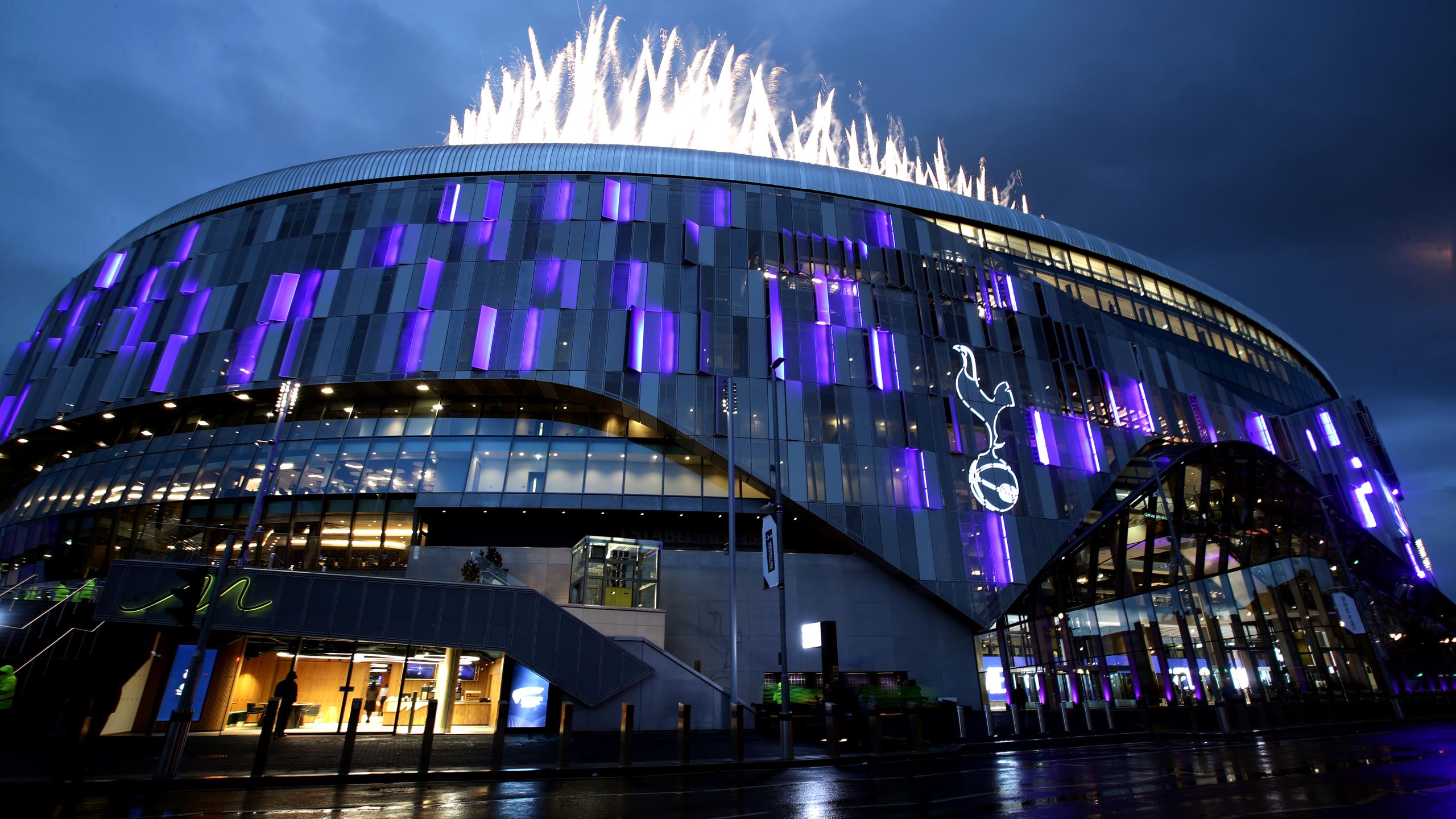 The iconic Tottenham Hotspur Stadium at night, illuminated in purple and with fireworks overhead.
