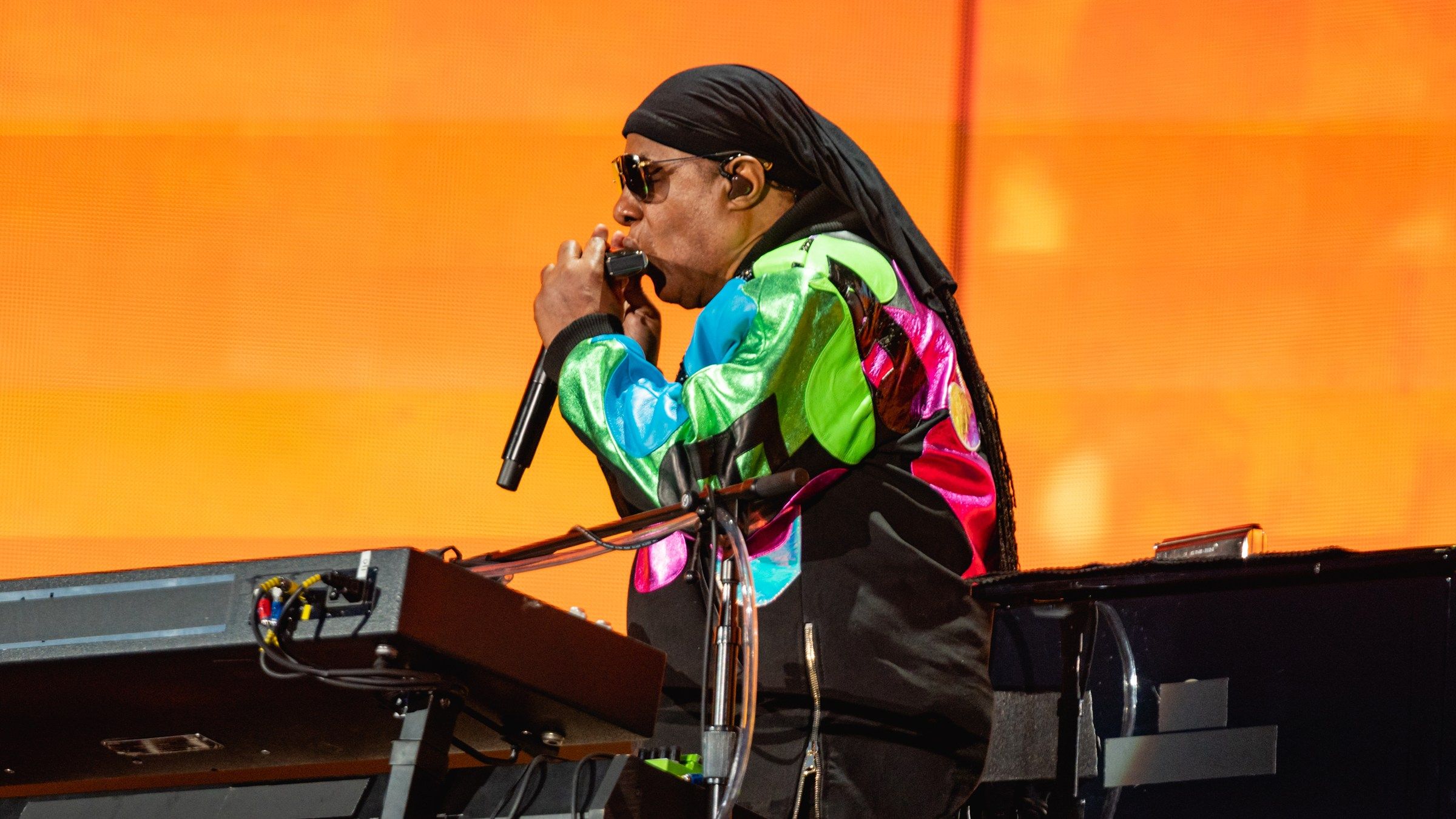Stevie Wonder playing the harmonica at his keyboard in front of an orange stage screen