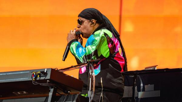 Stevie Wonder playing the harmonica at his keyboard in front of an orange stage screen