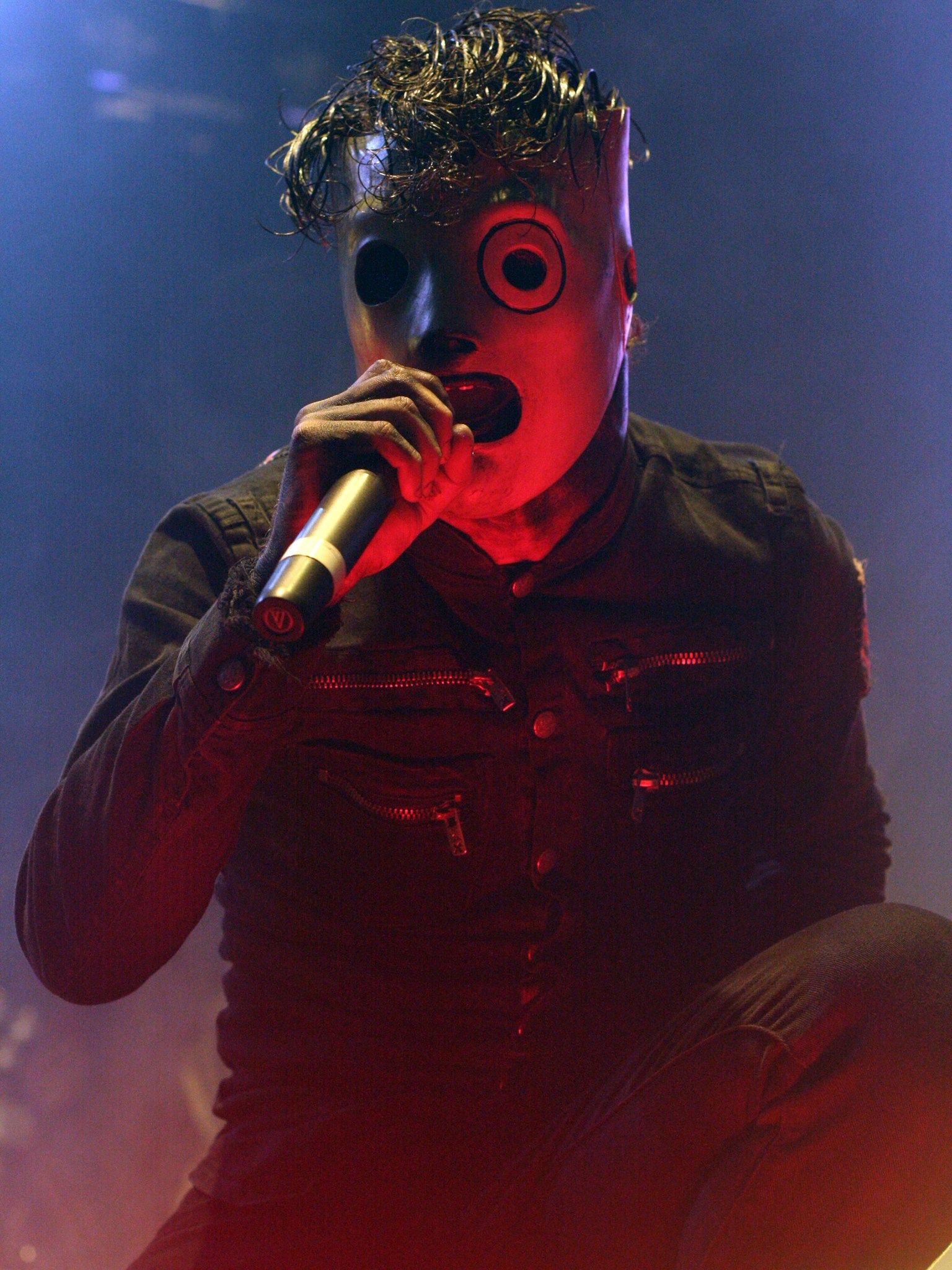 Corey Taylor of Slipknot singing into a microphone in his mask, lit red and blue