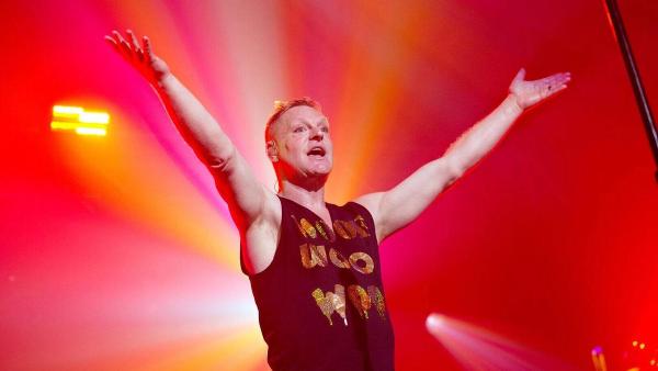 Andy Bell of Erasure singing on stage with both arms raised, lit by red and orange stage lights