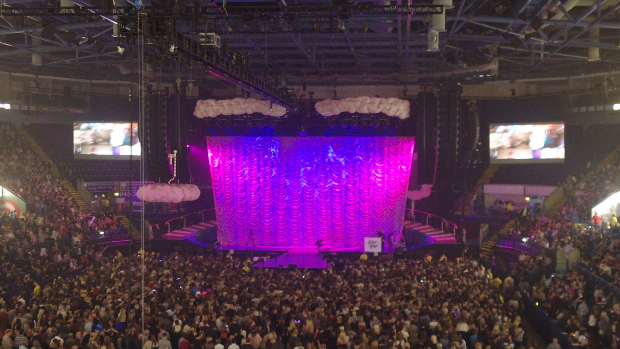 A packed arena floor and seating in Nottingham facing a curtained stage lit pink and purple before a concert