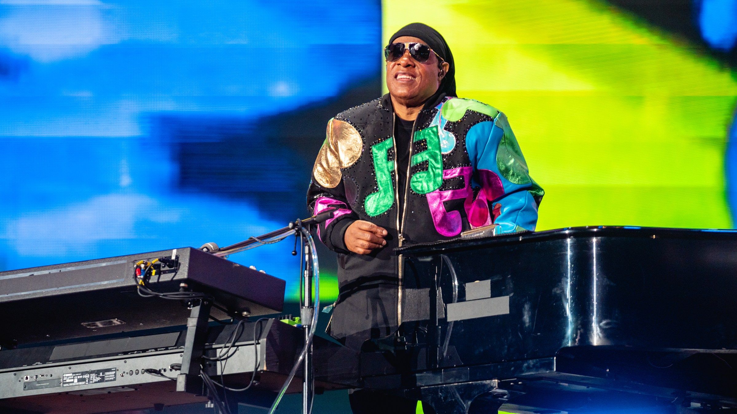 Stevie Wonder smiling between a keyboard and a grand piano on stage, with a blue and yellow video screen behind him