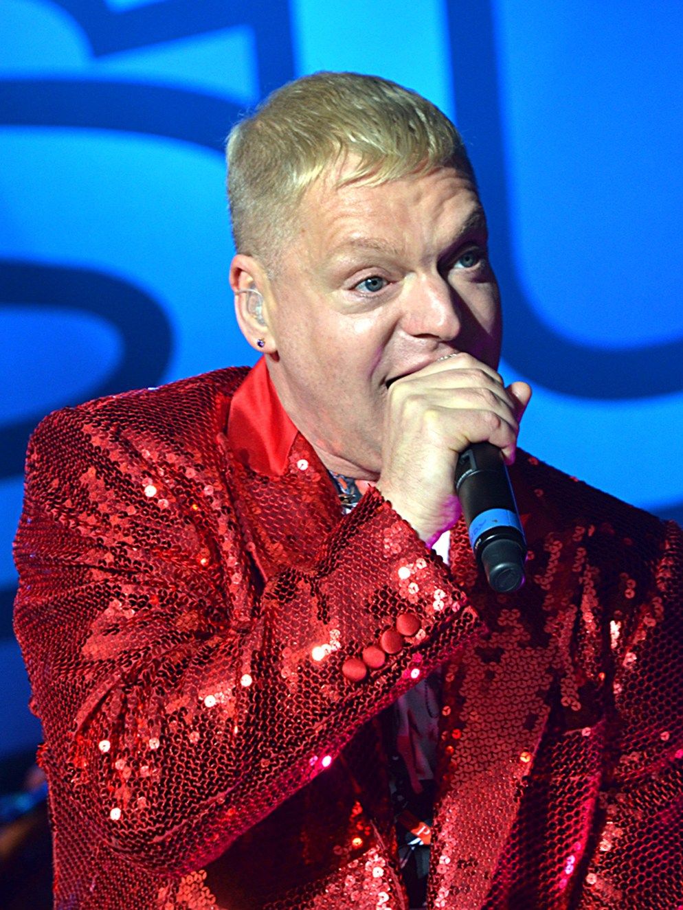 Andy Bell of Erasure singing in a red sequinned jacket against a bright blue backdrop