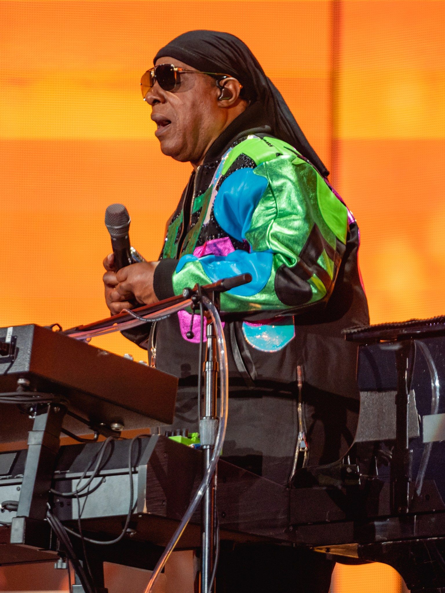 Stevie Wonder singing in profile at his keyboard, holding a microphone, against an orange stage backdrop
