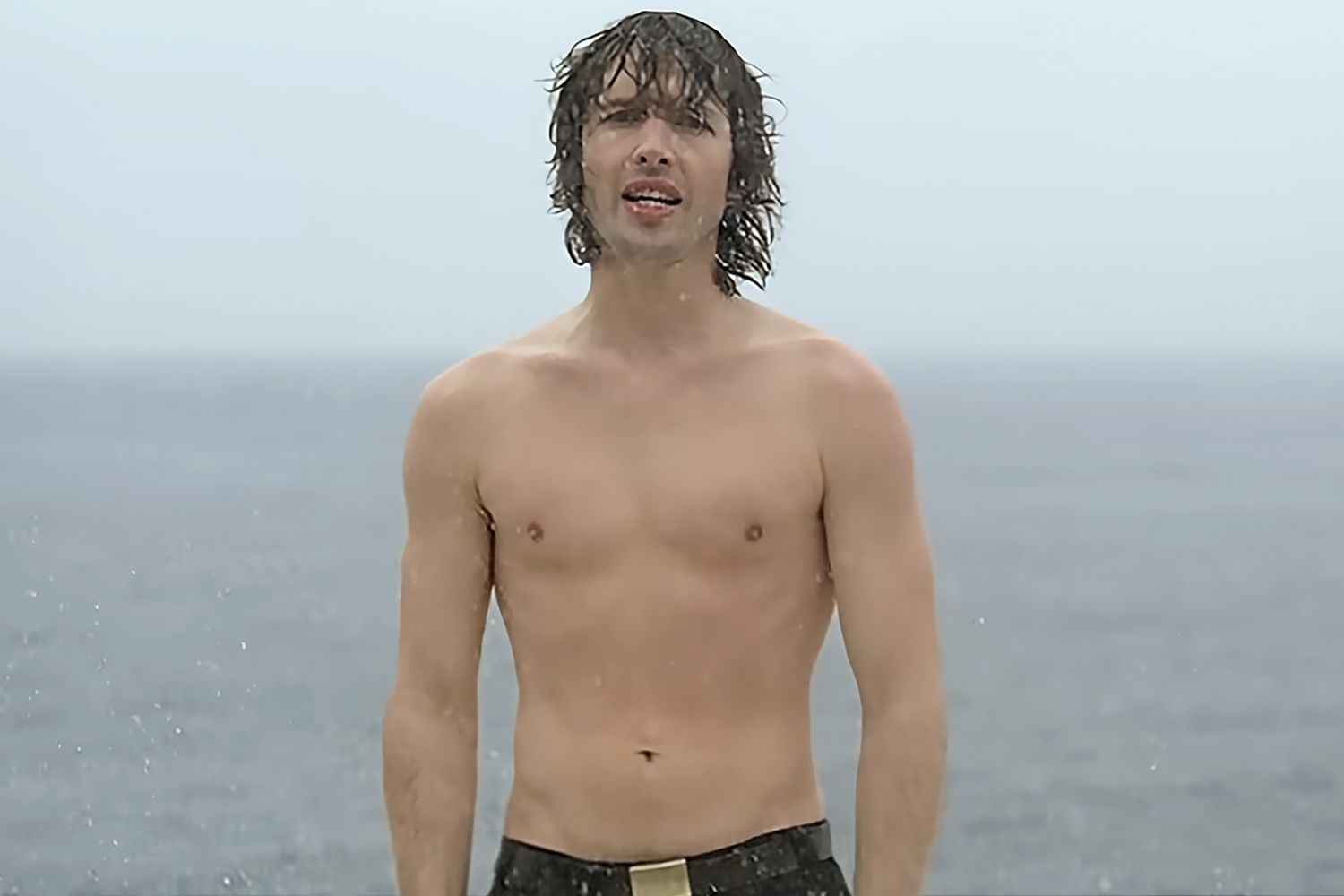 James Blunt shirtless with wet hair, singing in the rain with a grey sea behind him