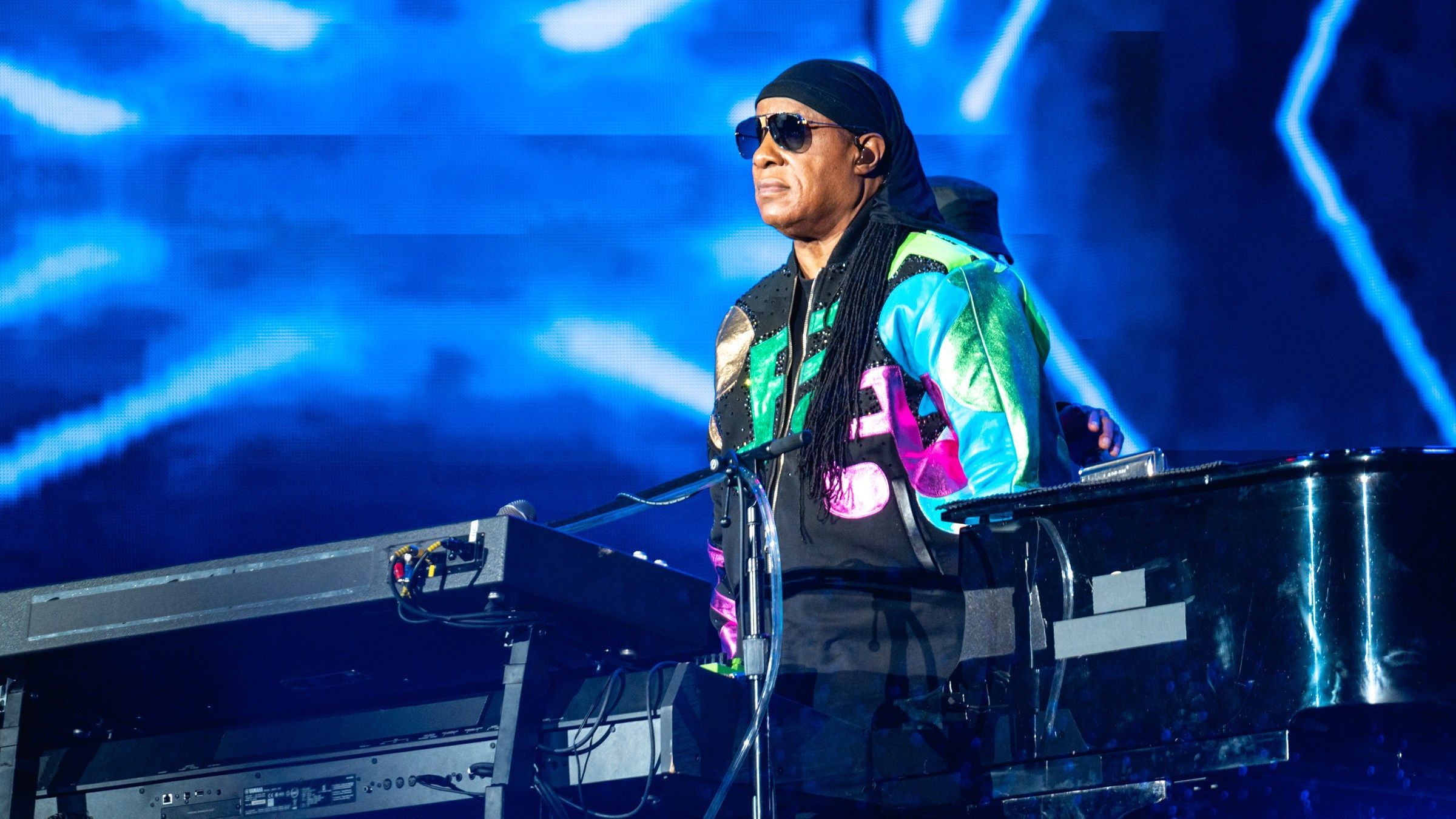 Stevie Wonder standing at his keyboard and grand piano in front of a blue video screen