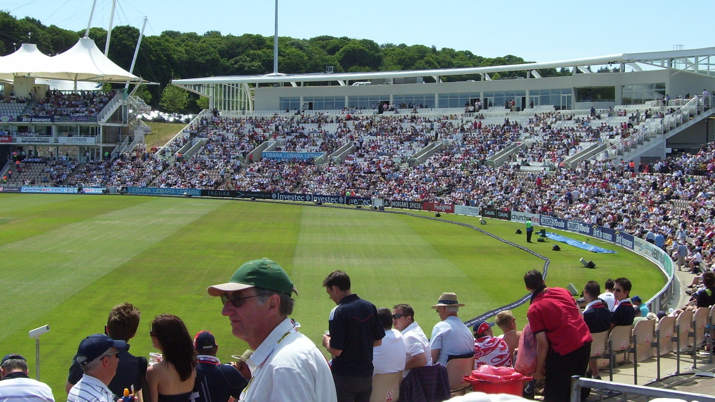 Spectators in summer sunshine in the stands at the Southampton cricket ground now called the Utilita Bowl, with the West Stand full and the outfield in front