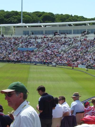 Spectators in summer sunshine in the stands at the Southampton cricket ground now called the Utilita Bowl, with the West Stand full and the outfield in front