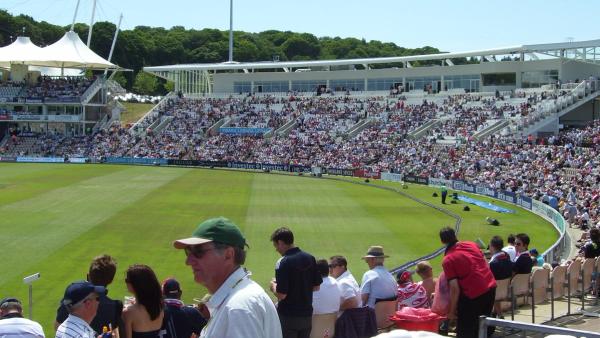 Spectators in summer sunshine in the stands at the Southampton cricket ground now called the Utilita Bowl, with the West Stand full and the outfield in front