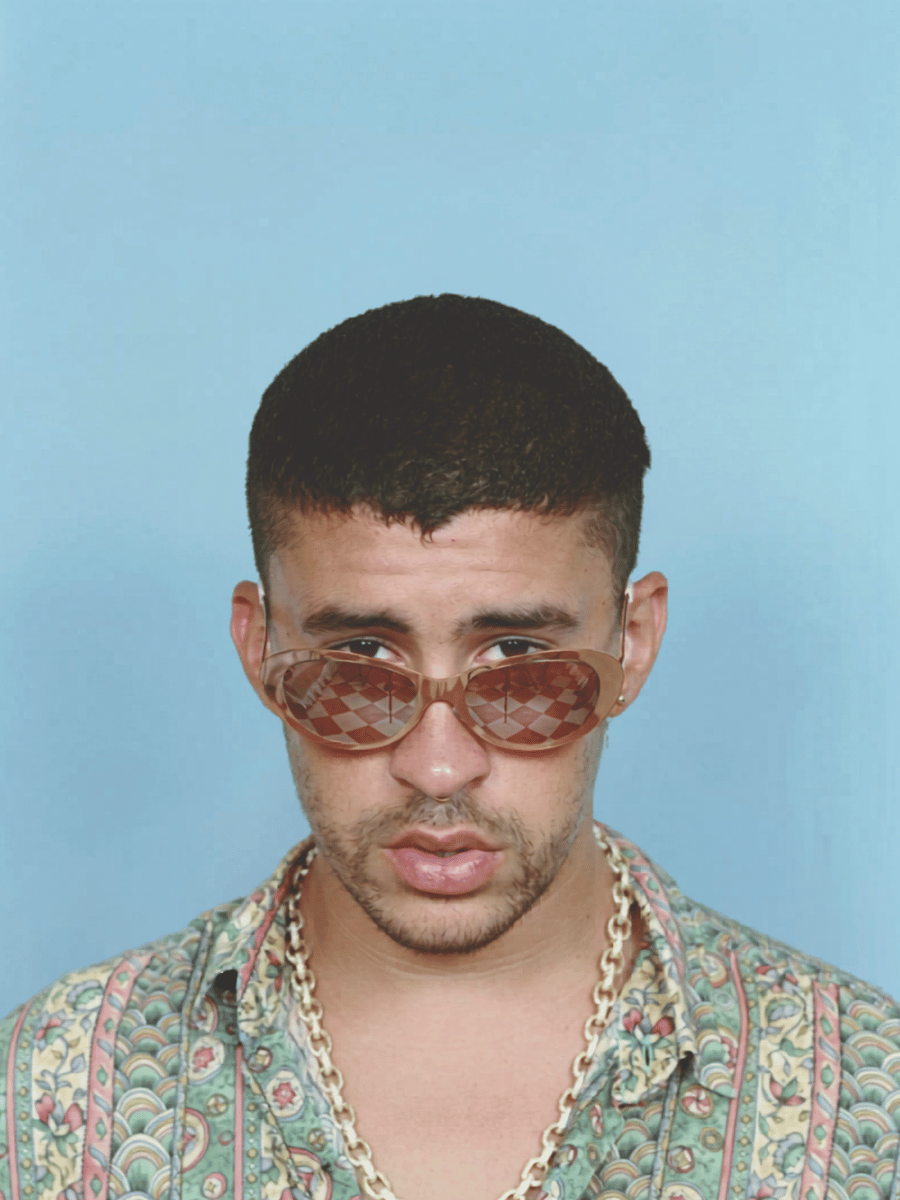 Bad Bunny in unique sunglasses, patterned shirt, and gold chain, studio portrait.