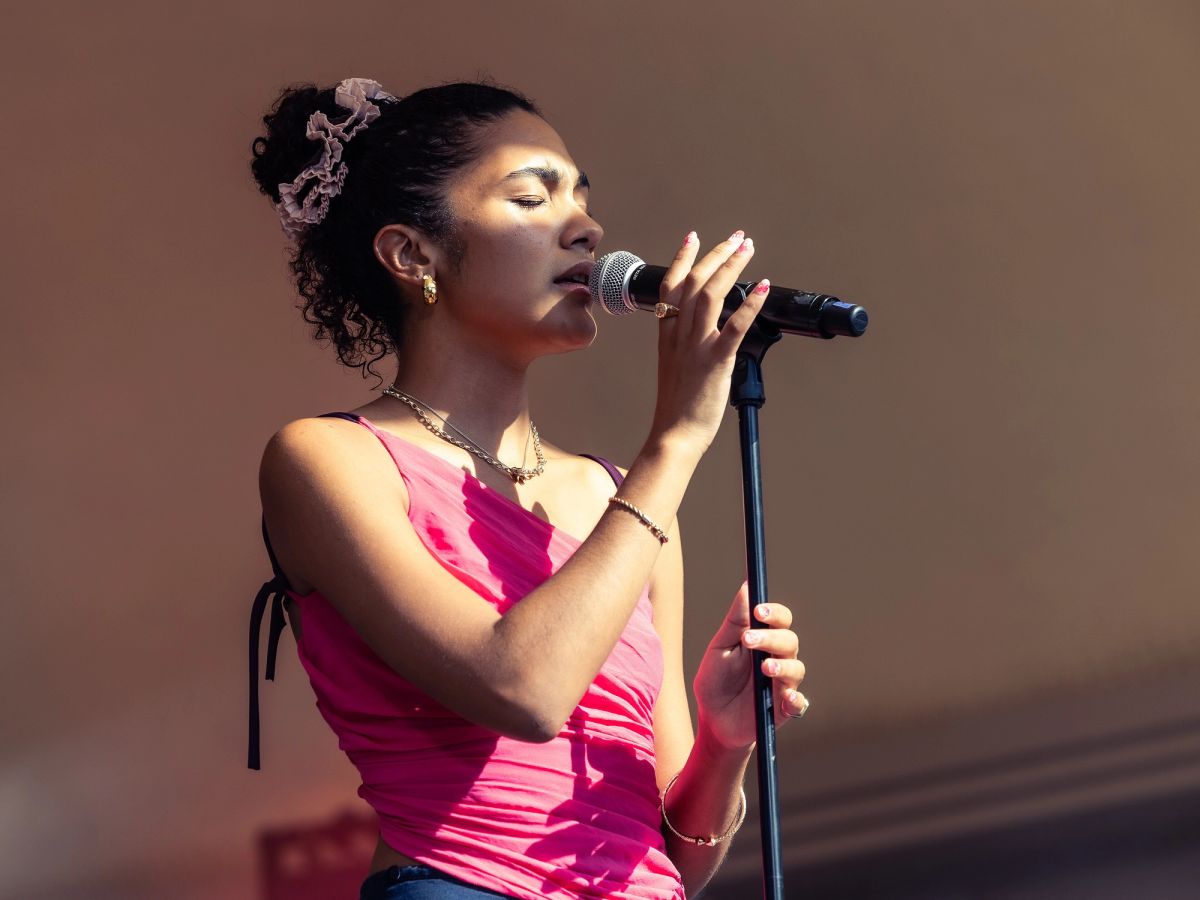 Olivia Dean singing into a microphone on a stand, eyes closed, wearing a pink top