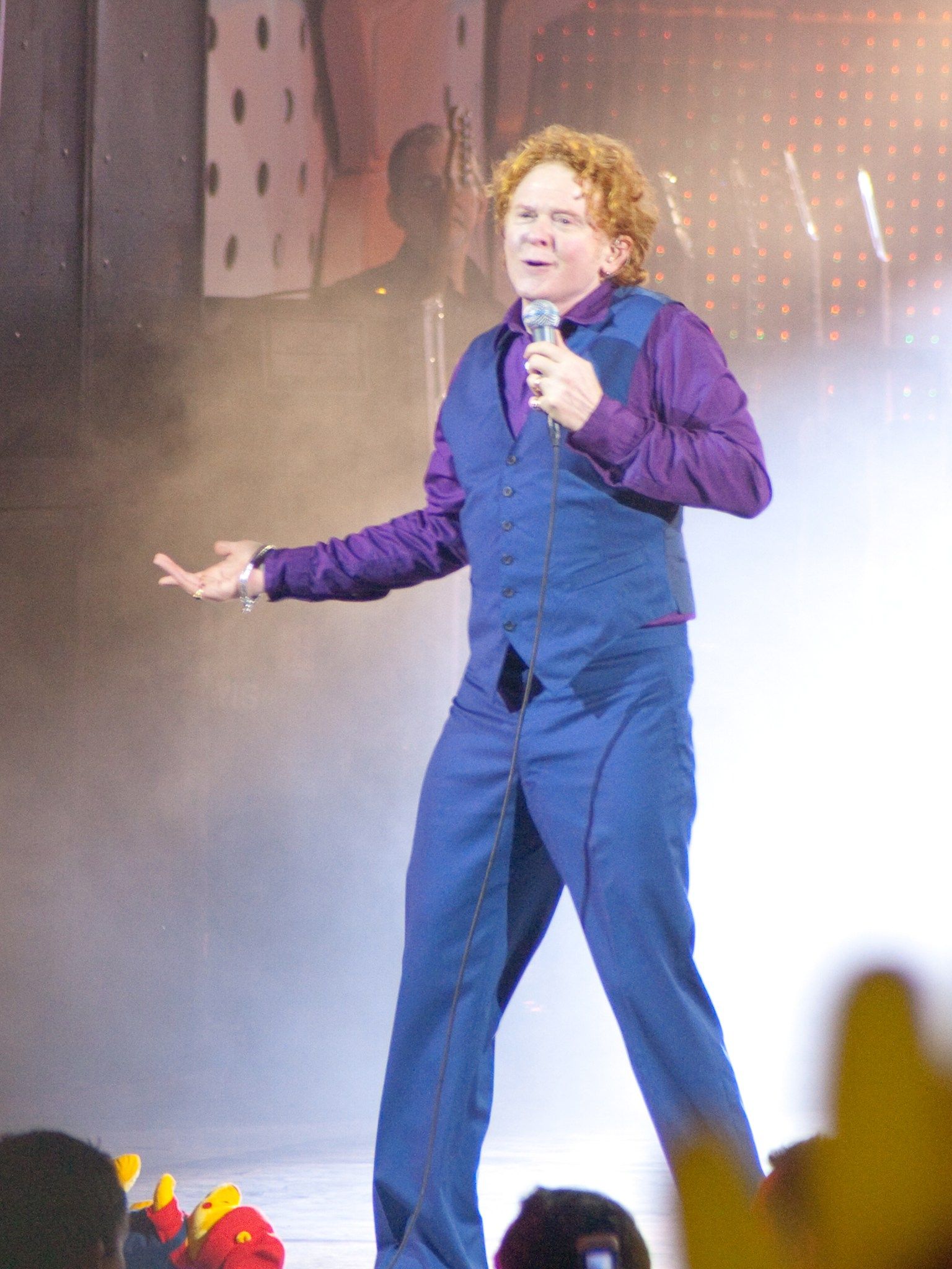 Mick Hucknall singing to the crowd through stage haze, wearing a blue waistcoat and purple shirt