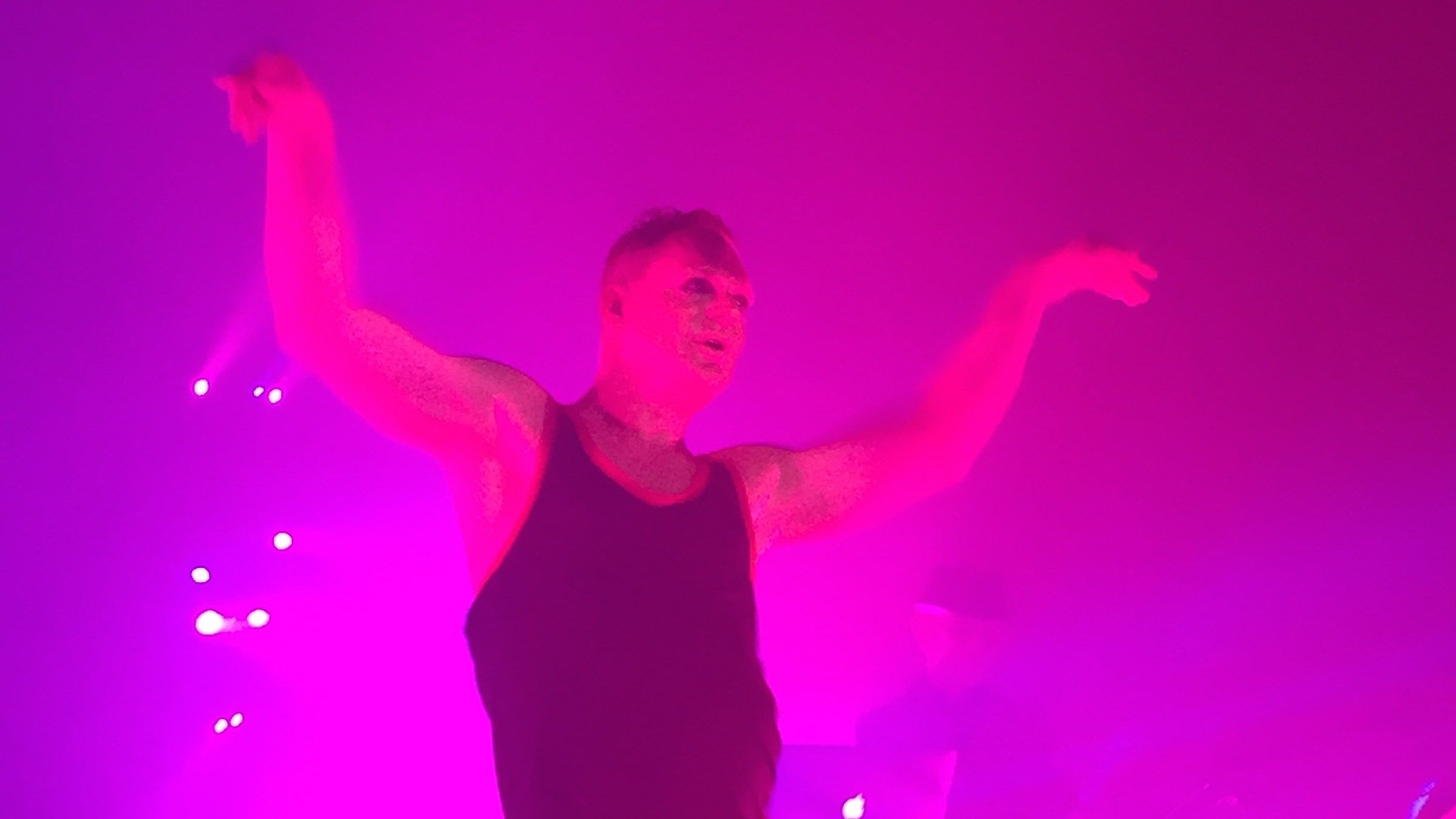 Andy Bell of Erasure dancing on stage with his arms spread, bathed in pink and purple stage light
