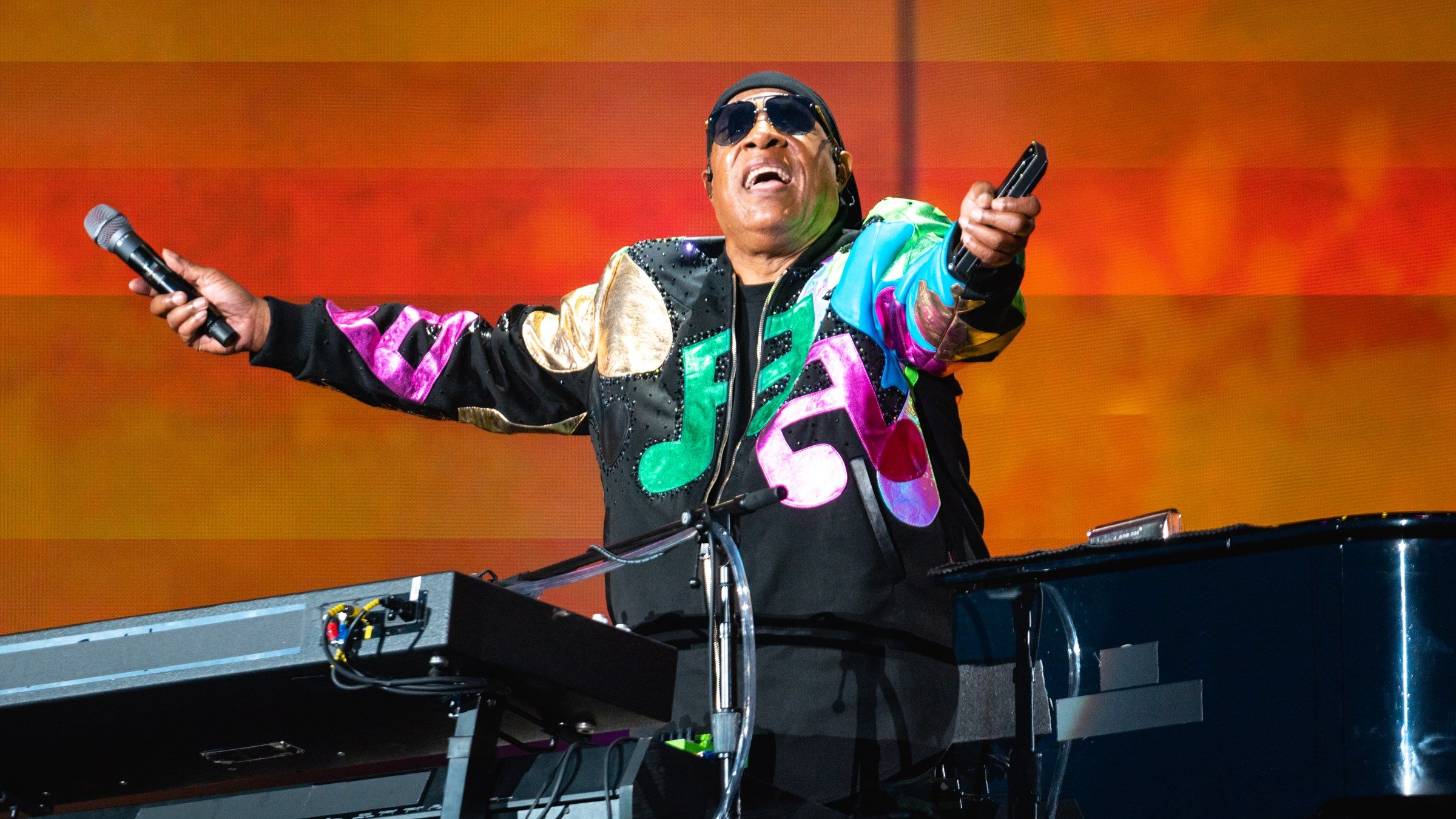 Stevie Wonder singing on stage with his arms outstretched, a microphone in one hand and a harmonica in the other, in front of an orange screen