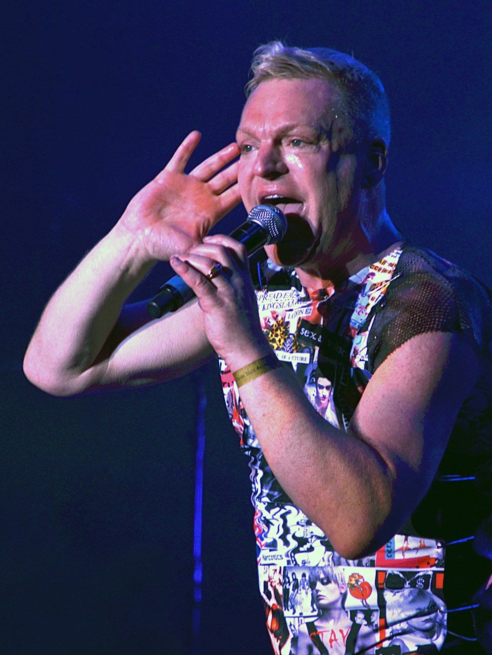 Andy Bell of Erasure singing into a microphone with one hand raised beside his face, against a dark blue background