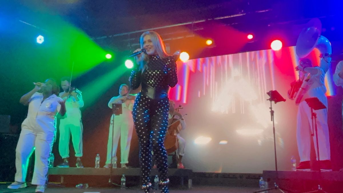 Becky Hill singing on stage under coloured lights, with string and brass players behind her