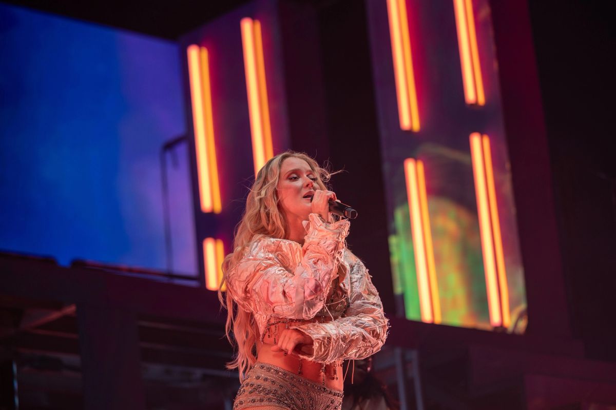 Zara Larsson singing on stage in a metallic jacket, with orange light panels behind her
